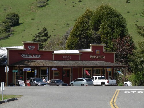 A quaint red general store with a sign reading "De Carly Emporium," surrounded by greenery and parked cars.