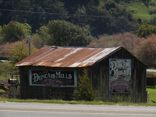 Rustic wooden building with a corrugated metal roof, featuring vintage signs for "Duncan's Mills" and "Goods and Groceries."