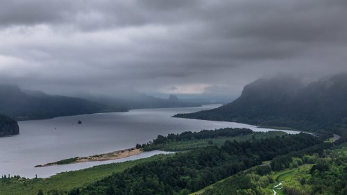 A misty river flows through lush green landscapes, surrounded by mountains under a cloudy sky.