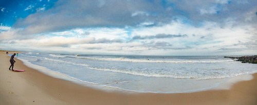 A surfer stands on the beach, preparing to enter the ocean under a cloudy sky with gentle waves.