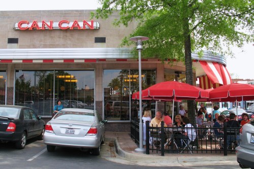 Exterior view of Can Can restaurant with outdoor seating, red umbrellas, and cars parked nearby.