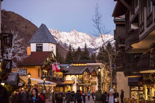 A charming village street adorned with lights, surrounded by mountains at dusk. People stroll past festive shops.