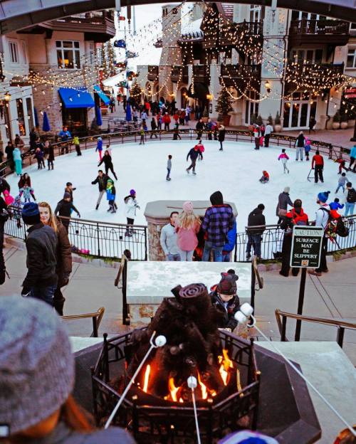A bustling ice skating rink surrounded by festive lights, with people skating and roasting marshmallows by a fire.