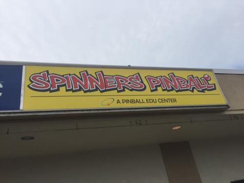 Sign for "Spinners Pinball," a pinball education center, featuring colorful lettering against a blue sky.