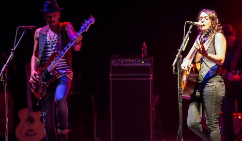 A male bassist and a female singer perform on stage, illuminated by colorful lights, with instruments in the background.