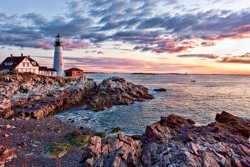 A scenic lighthouse on rocky shores at sunset, with colorful clouds and calm waters in the background.