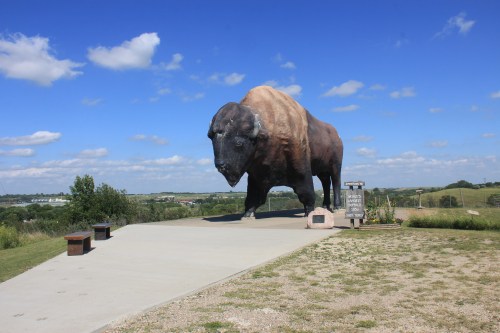 A large bison statue stands on a concrete platform, surrounded by green grass and a blue sky with fluffy clouds.