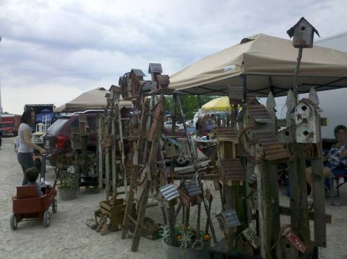 A craft market scene with unique birdhouses displayed under tents, and a child in a wagon nearby.