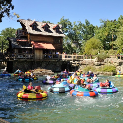 People enjoying a sunny day floating on colorful inner tubes in a river near a rustic wooden building.