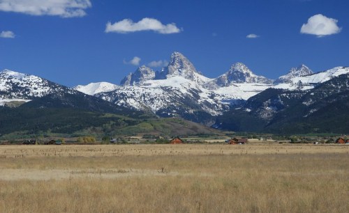 Snow-capped mountains rise majestically against a blue sky, with a grassy field and distant cabins in the foreground.