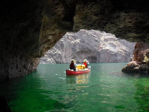 Two people in a red canoe paddle through a rocky cave, surrounded by green water and steep cliffs.