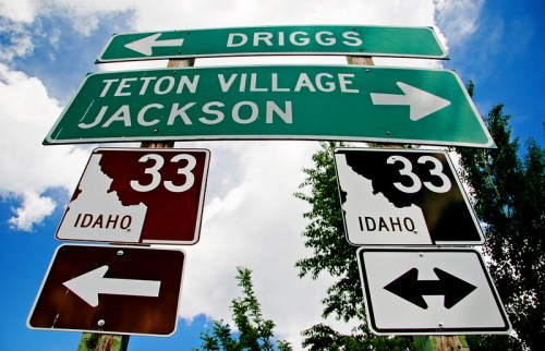 Road signs indicating directions to Driggs, Teton Village, and Jackson, with a blue sky and trees in the background.