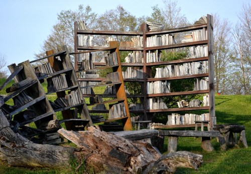 An artistic installation of weathered wooden bookshelves, partially collapsed, surrounded by greenery.