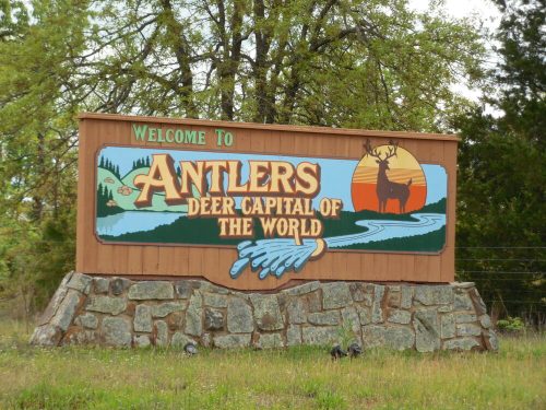 Welcome sign for Antlers, "Deer Capital of the World," featuring a deer, river, and scenic background.