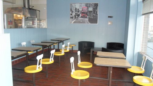 A small, empty café with yellow and black chairs, wooden tables, and a large photo of a double-decker bus on the wall.