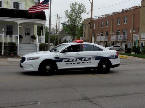 A police car with lights on, marked "Orland Hills," parked on a street near residential buildings.