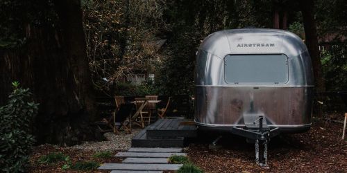 A shiny Airstream trailer parked in a wooded area, with a wooden deck and outdoor seating nearby.