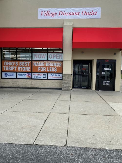 Exterior of Village Discount Outlet with a sign stating "Now Open" and promotions for thrift store deals.