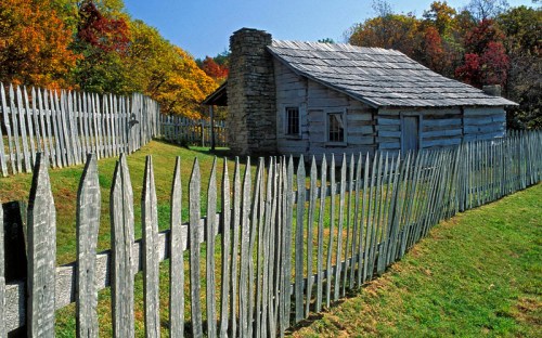A rustic log cabin surrounded by a wooden picket fence and colorful autumn trees in the background.