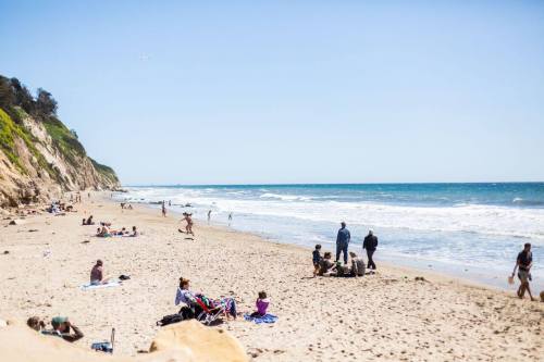 A sunny beach scene with people relaxing, playing, and walking along the shore, with waves gently lapping at the sand.