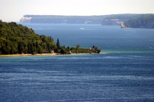 A serene view of a blue lake surrounded by lush green trees and distant cliffs under a clear sky.