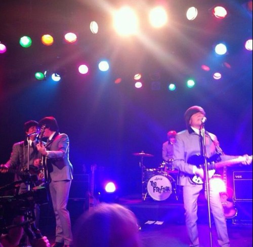 A band performs on stage under colorful lights, with musicians in suits and a drummer in the background.