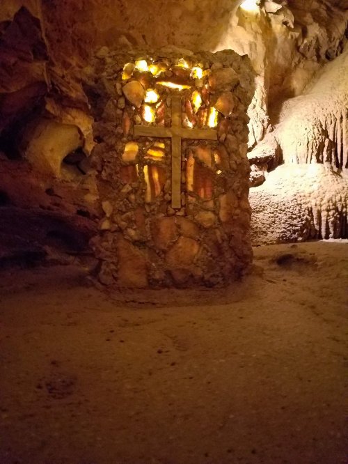 A stone cross illuminated inside a cave, surrounded by rock formations and soft lighting.