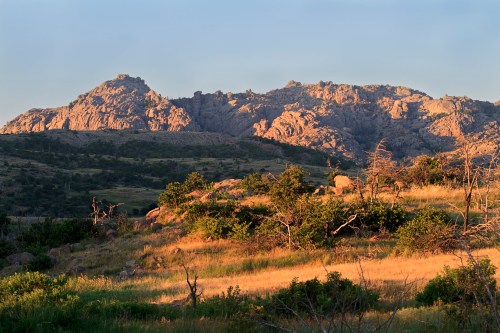 Sunlit rocky mountains rise above a grassy landscape, with scattered trees and a clear sky in the background.