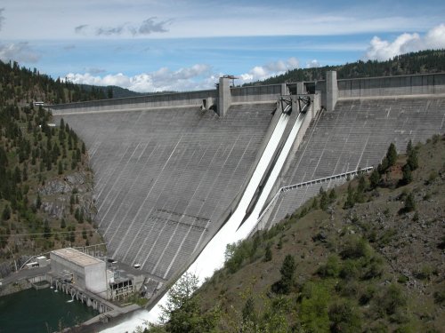 A large concrete dam with water cascading down its face, surrounded by green hills and a blue sky.