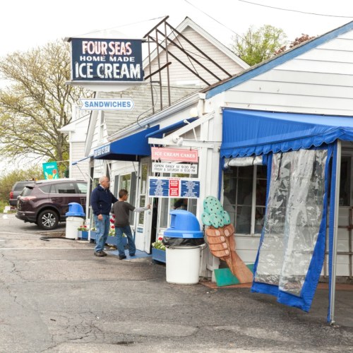 A quaint ice cream shop with a blue awning, featuring a sign for homemade ice cream and a customer at the window.