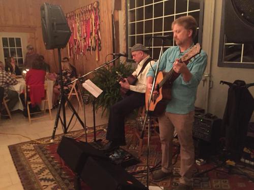 Two musicians perform on stage with guitars in a cozy venue, while guests enjoy dinner in the background.