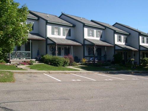 Row of two-story townhouses with porches, surrounded by greenery and a clear blue sky. Parking spaces in front.