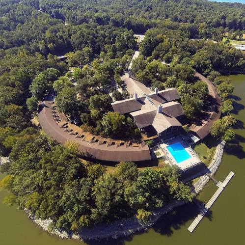 Aerial view of a resort surrounded by trees, featuring a pool and a winding building layout near a lake.