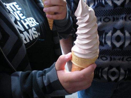 Two hands hold ice cream cones with swirled soft serve, one wearing a striped sweater and the other a graphic t-shirt.