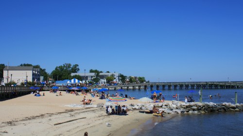 A sunny beach scene with people relaxing on the sand, umbrellas, and a pier extending into the water.