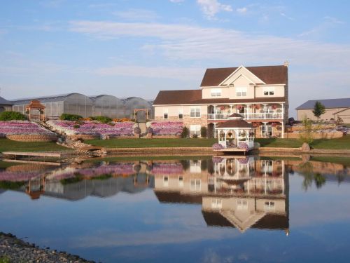 A picturesque house with flower gardens, reflecting in a calm pond under a clear blue sky. Greenhouses are in the background.
