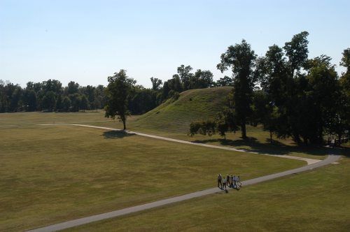 A group of people walks along a path in a grassy park with a large mound and trees in the background.