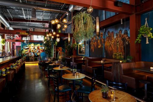A vibrant restaurant interior featuring colorful murals, hanging plants, and warm lighting with wooden tables and a bar.