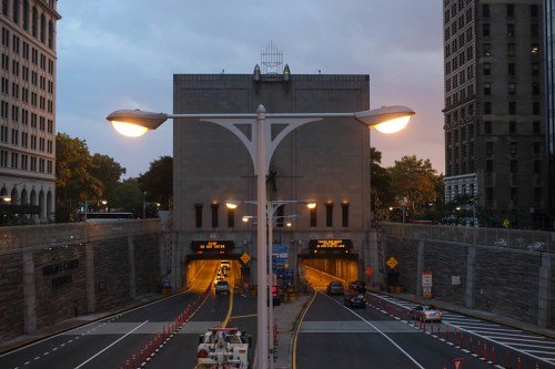 View of a city tunnel entrance with traffic lights and street lamps, surrounded by tall buildings at dusk.