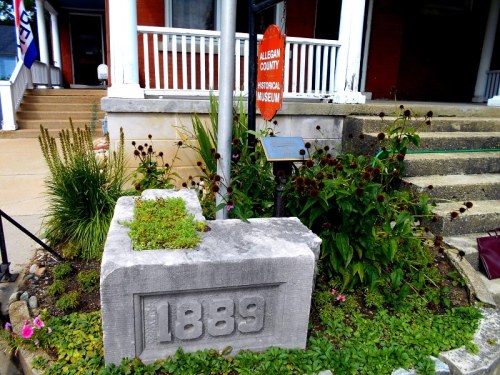 Stone marker with "1889" in front of a historical museum, surrounded by plants and a sign for the museum.