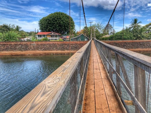 A wooden suspension bridge spans a calm river, with lush greenery and houses visible in the background under a blue sky.