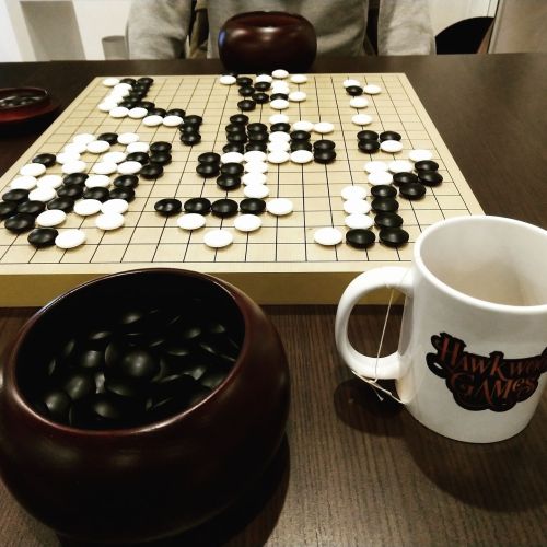 A Go board with black and white stones, a tea mug, and a bowl of stones on a wooden table.
