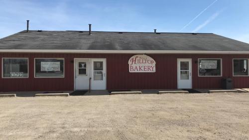A red building with a sign reading "Hilltop Bakery," featuring closed signs on the doors and a gravel parking area.