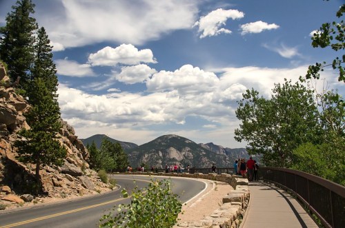 A scenic road curves through a mountainous landscape, with trees and clouds under a bright blue sky.