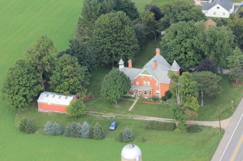 Aerial view of a large red house surrounded by trees, a barn, and a green lawn, with a road nearby.