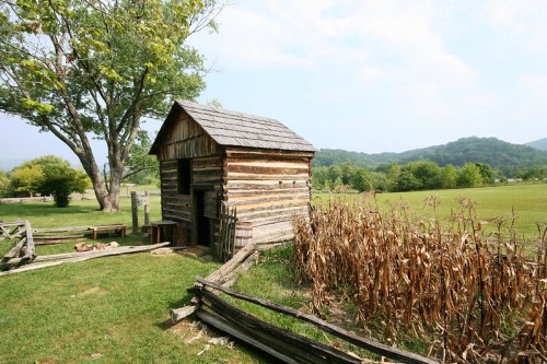 A rustic wooden cabin beside a field of dried corn, with rolling hills and trees in the background under a blue sky.