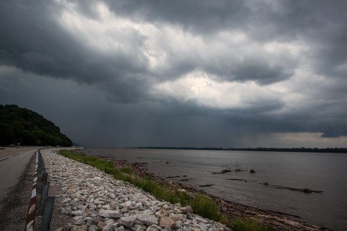A stormy sky looms over a river, with dark clouds and debris along the water's edge beside a road.