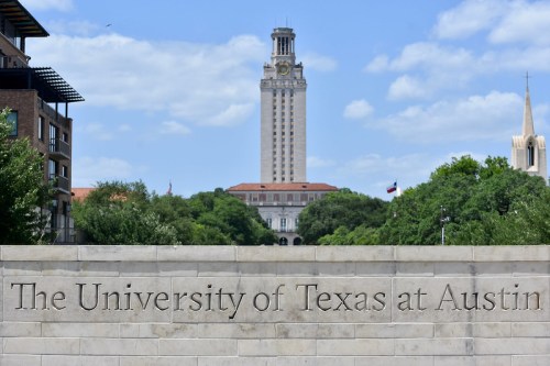 Sign for the University of Texas at Austin with the iconic tower in the background against a blue sky.