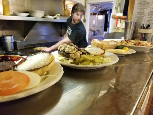 A chef prepares food in a kitchen, with plates of burgers and toppings lined up on the counter in the foreground.