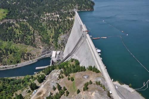 Aerial view of a large dam beside a lake, surrounded by green hills and trees.